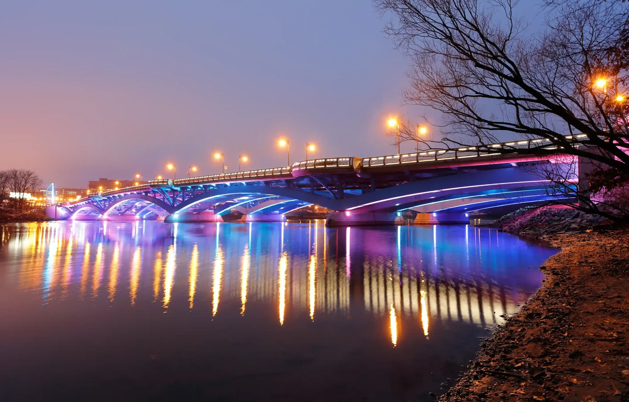 A large bridge over a river lit up from beneath with multi colored light